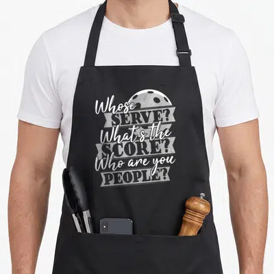 Score to serve man apron - Stacked white and grey text asking Whose Serve over a pickleball graphic — shown on a black cotton canvas apron against a white background