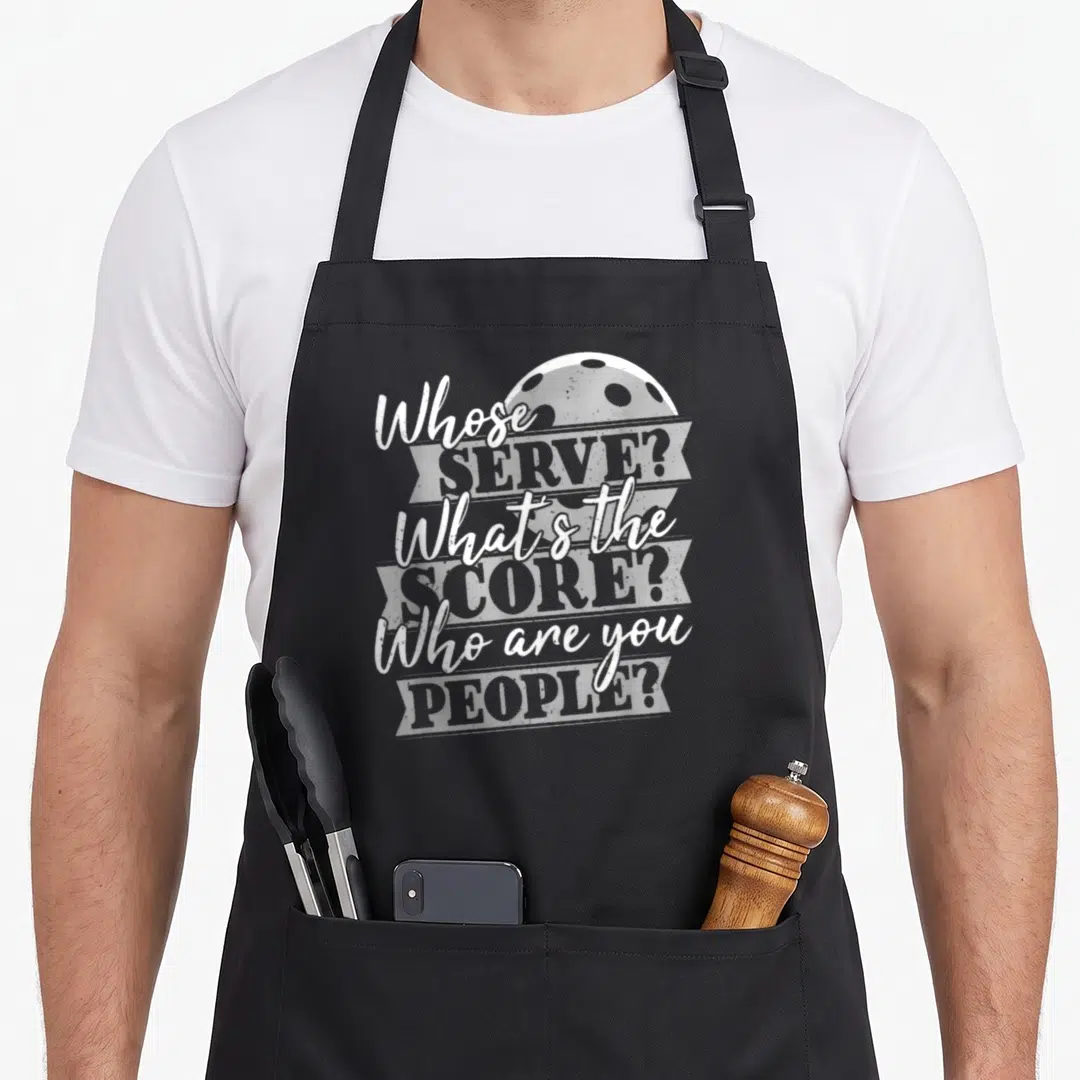 Score to serve man apron - Stacked white and grey text asking Whose Serve over a pickleball graphic — shown on a black cotton canvas apron against a white background