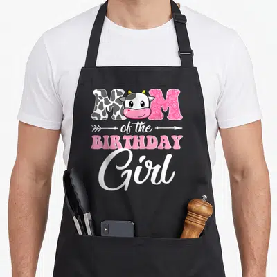 Farm mother cow print apron - MOM of the BIRTHDAY Girl with cartoon cow head in O, pink patterned letters, white arrow — shown on a black cotton canvas apron against a white background