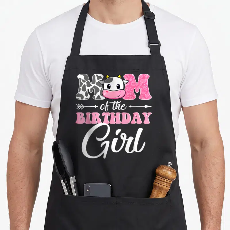 Farm mother cow print apron - MOM of the BIRTHDAY Girl with cartoon cow head in O, pink patterned letters, white arrow — shown on a black cotton canvas apron against a white background