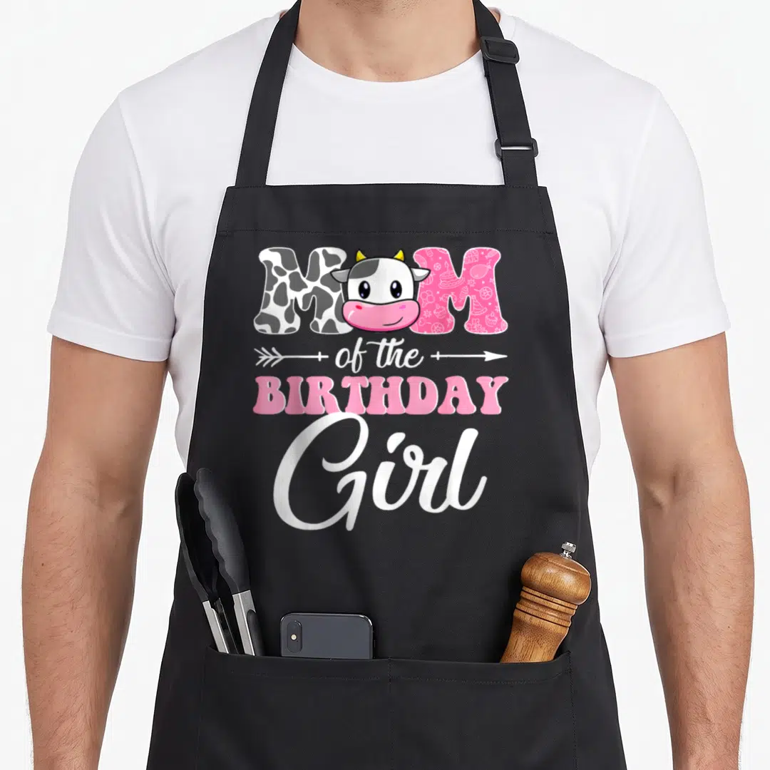 Farm mother cow print apron - MOM of the BIRTHDAY Girl with cartoon cow head in O, pink patterned letters, white arrow — shown on a black cotton canvas apron against a white background