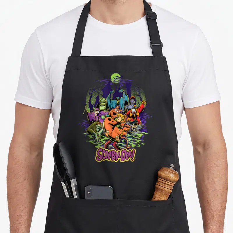 Shaggy decade apron - Cartoon Scooby-Doo and Shaggy running from monsters like a clown and knight near a purple castle — shown on a black cotton canvas apron against a white background
