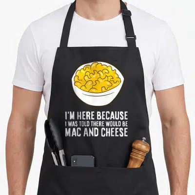 Would mac apron - Cartoon yellow macaroni bowl with white text reading I'm here because I was told there would be mac — shown on a black cotton canvas apron against a white background