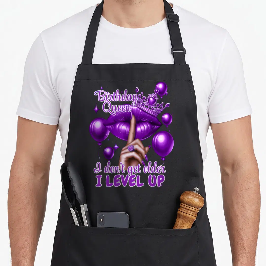 Older queen apron - Purple lips with crown, hand pointing up, balloons, and text reading Birthday Queen I Level Up — shown on a black cotton canvas apron against a white background