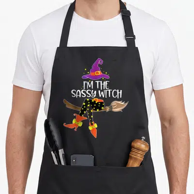 Sassy halloween witch apron - I'M THE SASSY WITCH text below purple hat and starry witch legs on broom — shown on a black cotton canvas apron against a white background