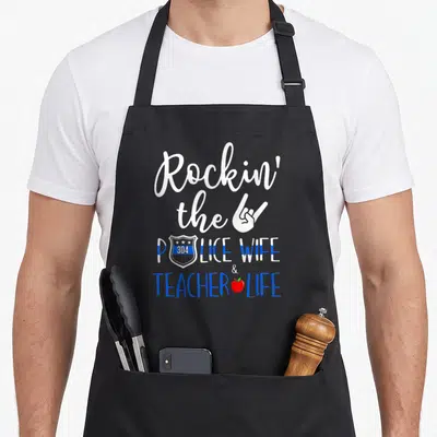 Rockin best teacher apron - Stacked text Rockin the Police Wife Teacher Life with hand gesture badge and red apple — shown on a black cotton canvas apron against a white background