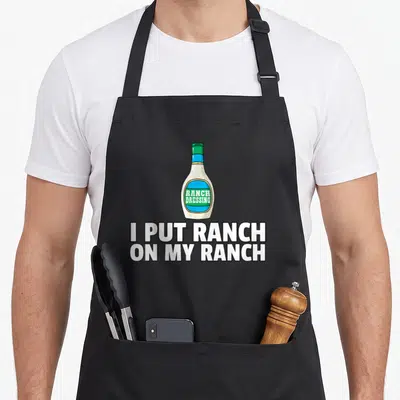 Sauce dressing apron - Cartoon ranch dressing bottle above I PUT RANCH ON MY RANCH text in white blue and green — shown on a black cotton canvas apron against a white background