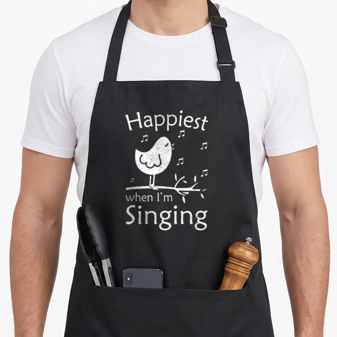 Sing singing bird apron - Distressed white bird perched on branch with Happiest when I'm Singing text — shown on a black cotton canvas apron against a white background