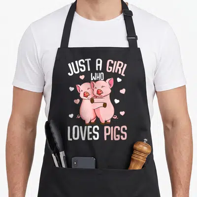 Vintage pig lover apron - Two pink cartoon pigs hugging with scattered hearts and text JUST A GIRL WHO LOVES PIGS — shown on a black cotton canvas apron against a white background