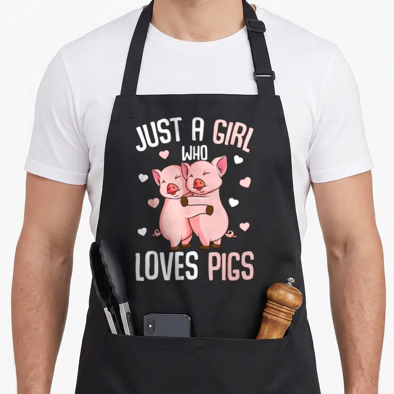 Vintage pig lover apron - Two pink cartoon pigs hugging with scattered hearts and text JUST A GIRL WHO LOVES PIGS — shown on a black cotton canvas apron against a white background