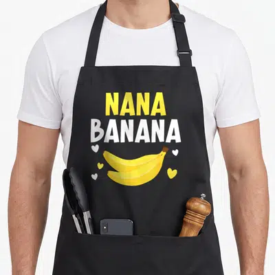 Nana banana grandma apron - Yellow NANA and white BANANA text above two bananas and five scattered hearts — shown on a black cotton canvas apron against a white background