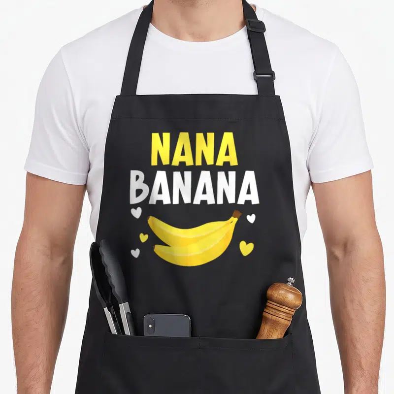 Nana banana grandma apron - Yellow NANA and white BANANA text above two bananas and five scattered hearts — shown on a black cotton canvas apron against a white background