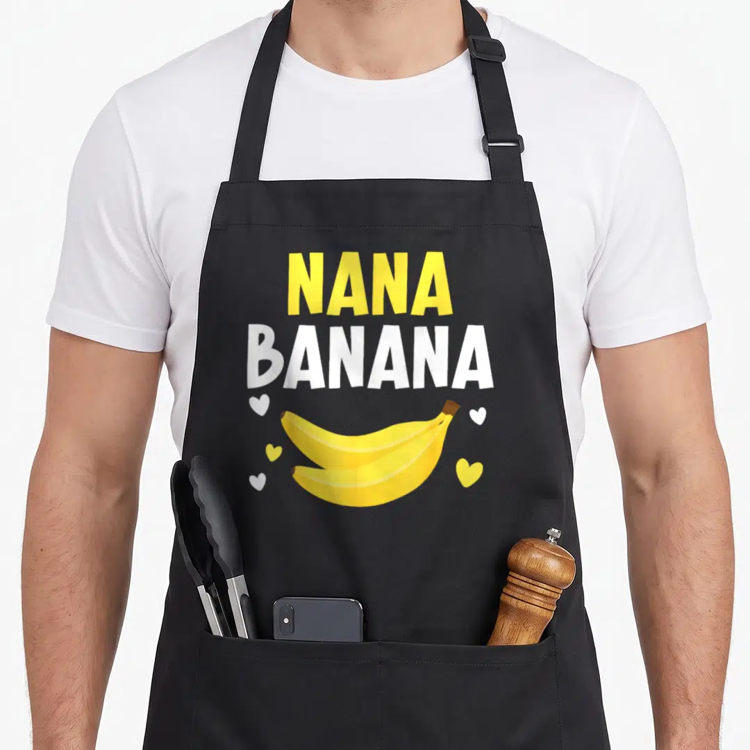 Nana banana grandma apron - Yellow NANA and white BANANA text above two bananas and five scattered hearts — shown on a black cotton canvas apron against a white background