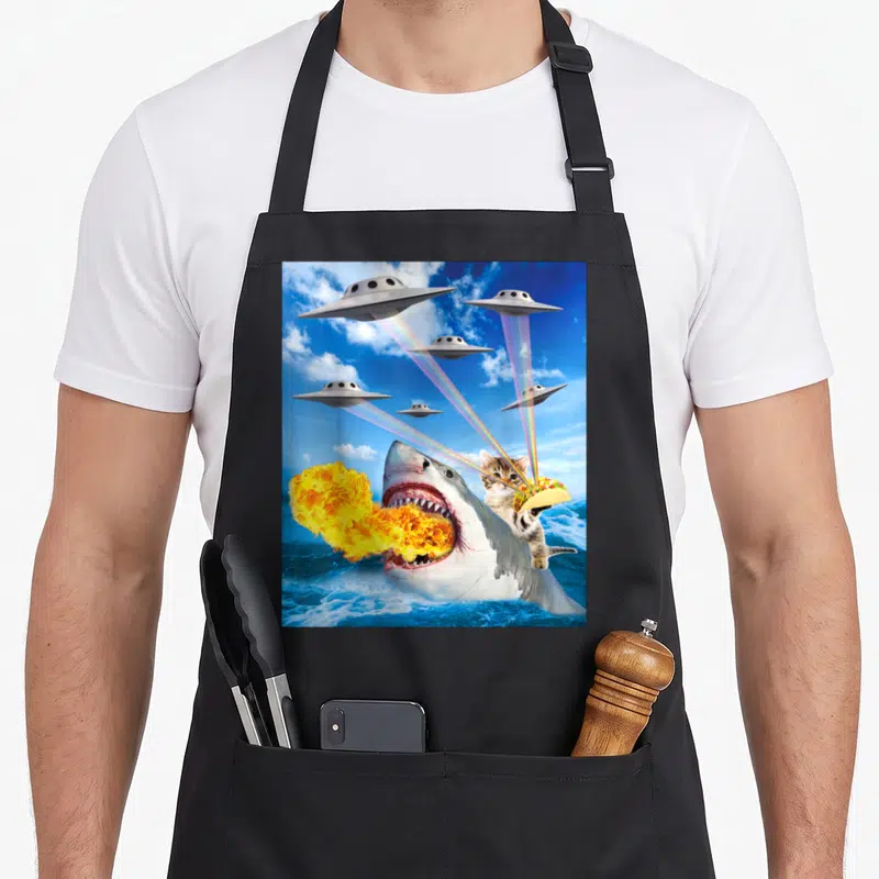 Riding cat apron - Shark breathing fire with kitten holding taco under six UFOs firing rainbow beams — shown on a black cotton canvas apron against a white background