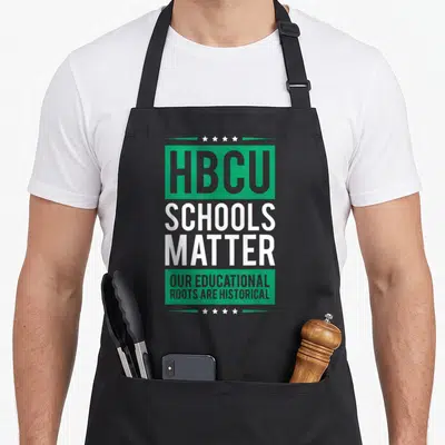 Hbcu college apron - HBCU SCHOOLS MATTER text in green and white with eight stars in arches above and below — shown on a black cotton canvas apron against a white background