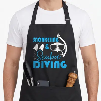 Snorkeling holiday apron - Blue stacked text reading SNORKELING & Scuba DIVING with white flippers mask and bubbles — shown on a black cotton canvas apron against a white background