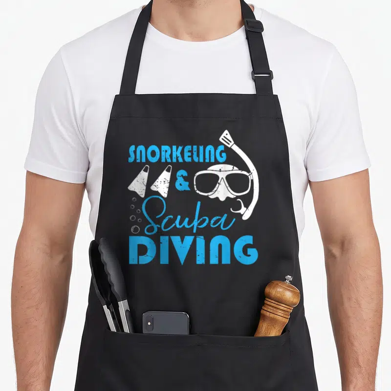 Snorkeling holiday apron - Blue stacked text reading SNORKELING & Scuba DIVING with white flippers mask and bubbles — shown on a black cotton canvas apron against a white background