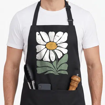 Boho gardener apron - Large white daisy with yellow center and green leaves in distressed retro style on black — shown on a black cotton canvas apron against a white background