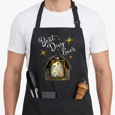 Manger womens christmas apron - Best Day Ever text above nativity scene in stable with yellow stars — shown on a black cotton canvas apron against a white background