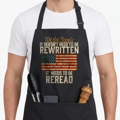 Rewritten novelty apron - Distressed USA flag with We the People text and It Needs to be Reread slogan on black — shown on a black cotton canvas apron against a white background