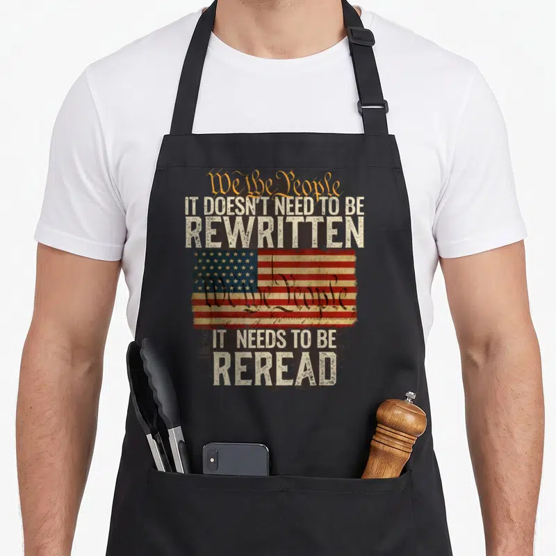 Rewritten novelty apron - Distressed USA flag with We the People text and It Needs to be Reread slogan on black — shown on a black cotton canvas apron against a white background