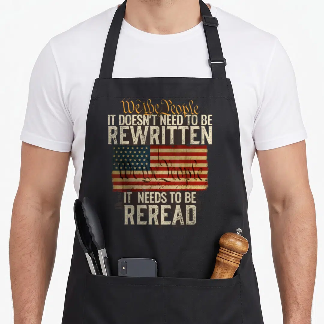 Rewritten novelty apron - Distressed USA flag with We the People text and It Needs to be Reread slogan on black — shown on a black cotton canvas apron against a white background