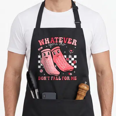 Valentine fall apron - Two pink cartoon socks on checkerboard with medical icons and text whatever you do don't fall — shown on a black cotton canvas apron against a white background
