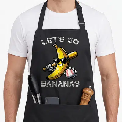 Youth banana apron - Cartoon banana with sunglasses holding a bat and baseball under white text — shown on a black cotton canvas apron against a white background