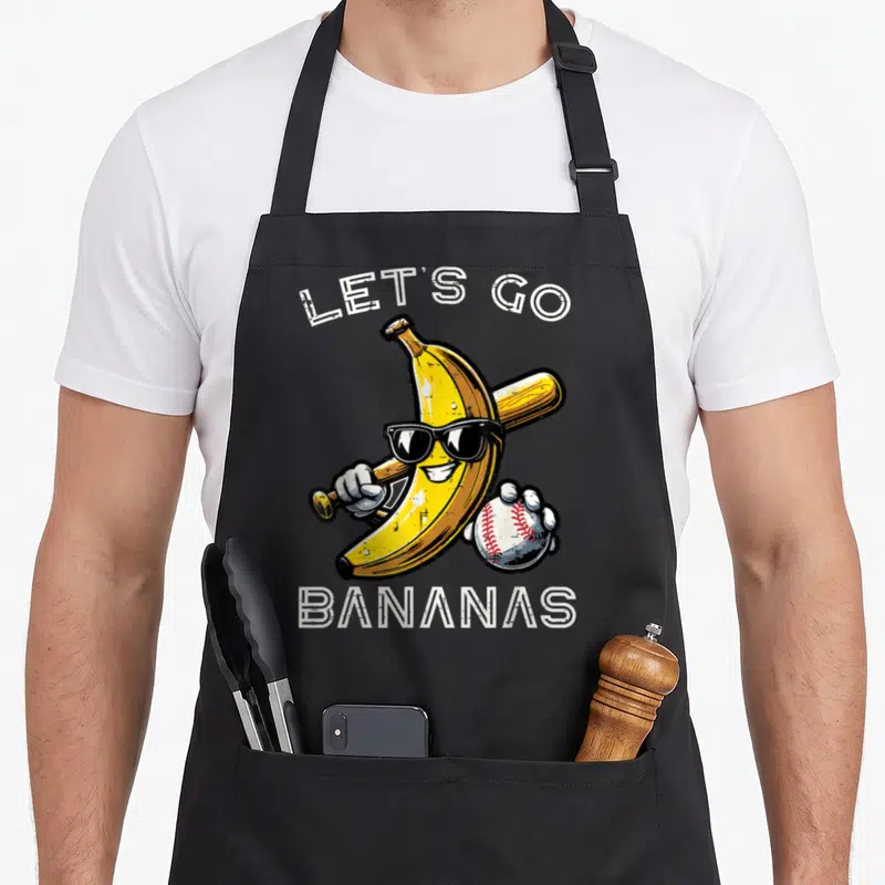 Youth banana apron - Cartoon banana with sunglasses holding a bat and baseball under white text — shown on a black cotton canvas apron against a white background