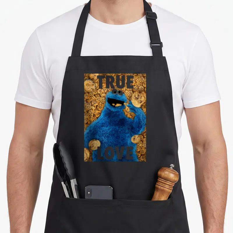 Beautiful cookies apron - Blue furry monster eating cookie surrounded by biscuits with TRUE LOVE text — shown on a black cotton canvas apron against a white background