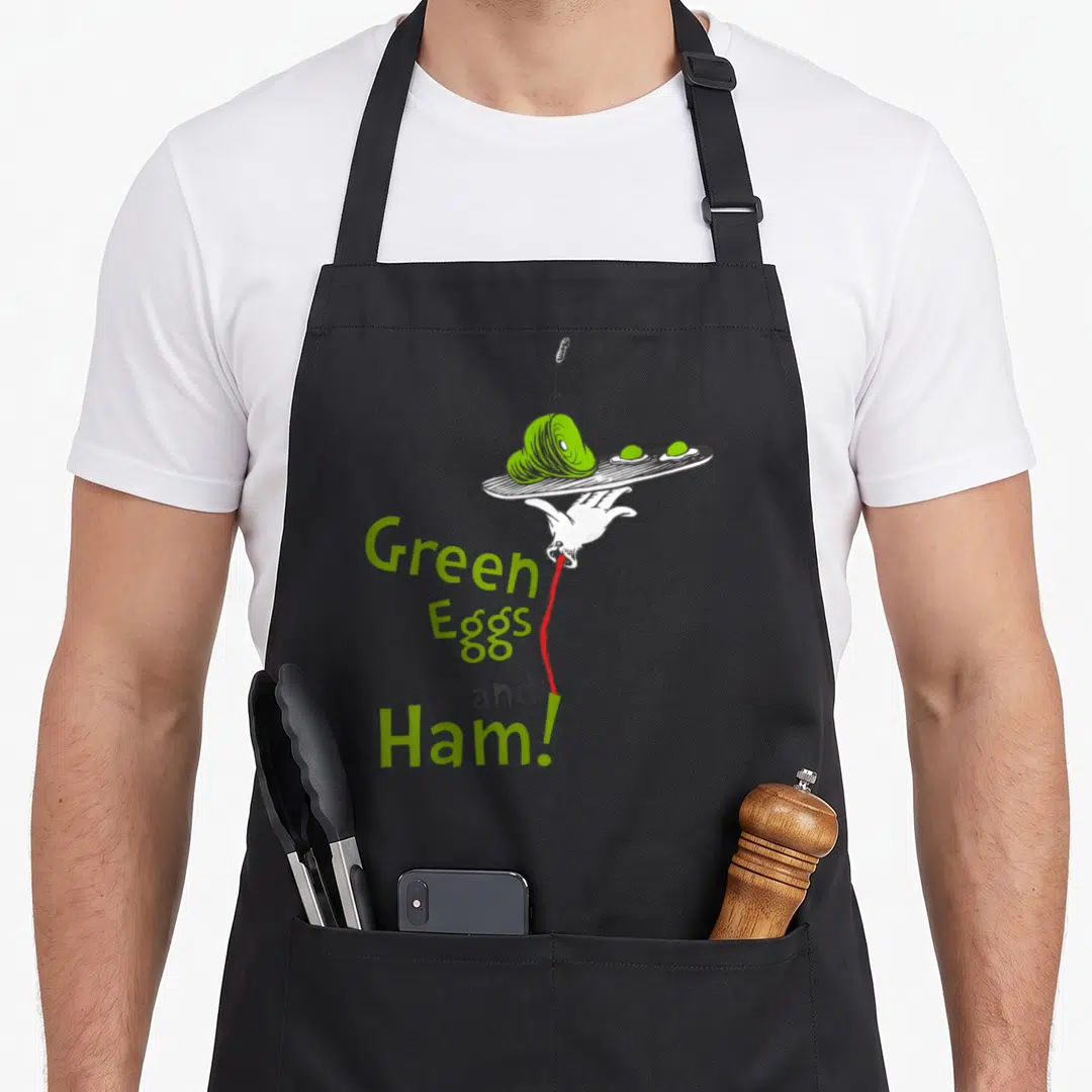 Ham egg apron - Hand holding plate of green ham and eggs with stacked text — shown on a black cotton canvas apron against a white background