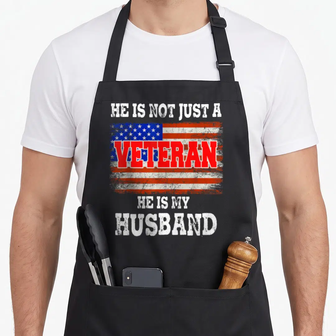 Flag best husband apron - Distressed USA flag with stacked text HE IS NOT JUST A VETERAN HE IS MY HUSBAND in red — shown on a black cotton canvas apron against a white background