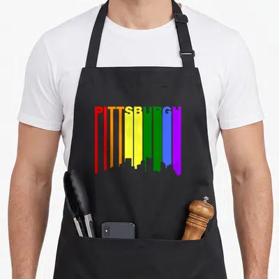 Pittsburgh gay pride apron - PITTSBURGH text with rainbow vertical stripes forming city skyline silhouette on black — shown on a black cotton canvas apron against a white background
