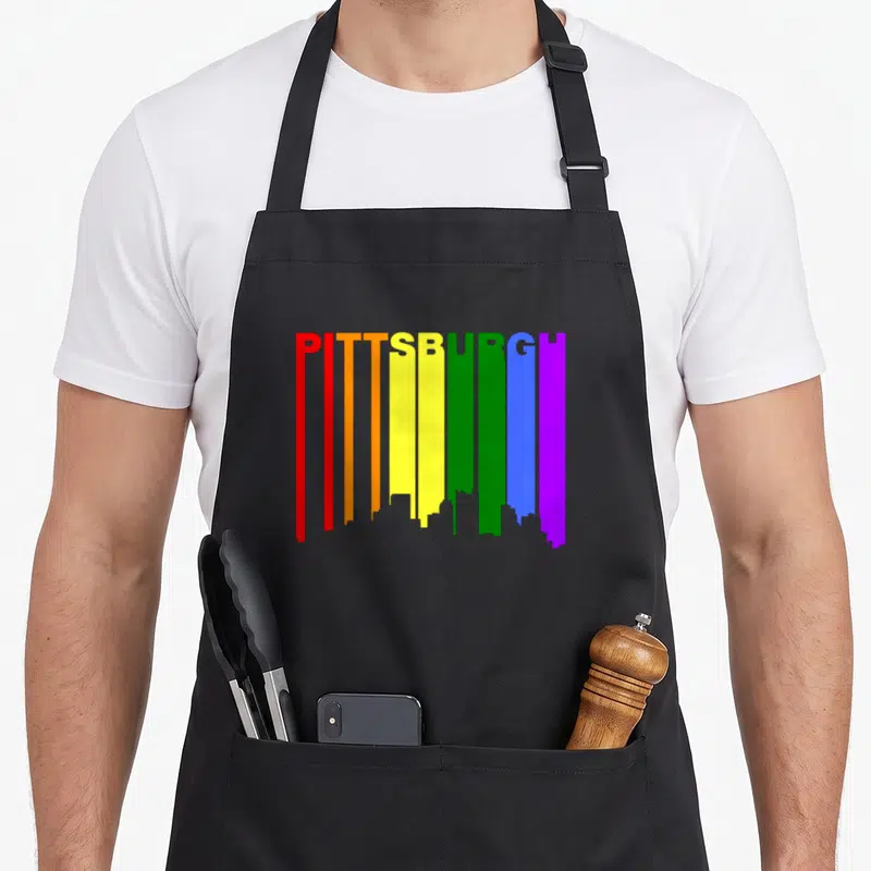 Pittsburgh gay pride apron - PITTSBURGH text with rainbow vertical stripes forming city skyline silhouette on black — shown on a black cotton canvas apron against a white background