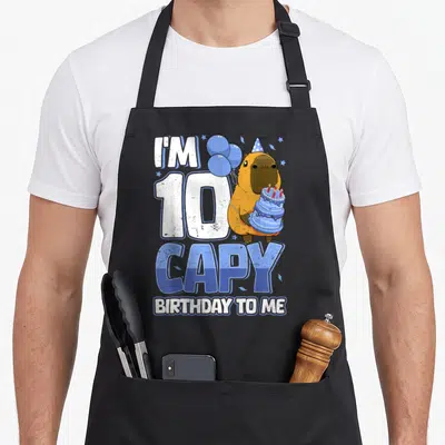 10th capybara birthday apron - Capybara in party hat holding blue cake with balloons and text I'M 10 CAPY BIRTHDAY TO ME — shown on a black cotton canvas apron against a white background