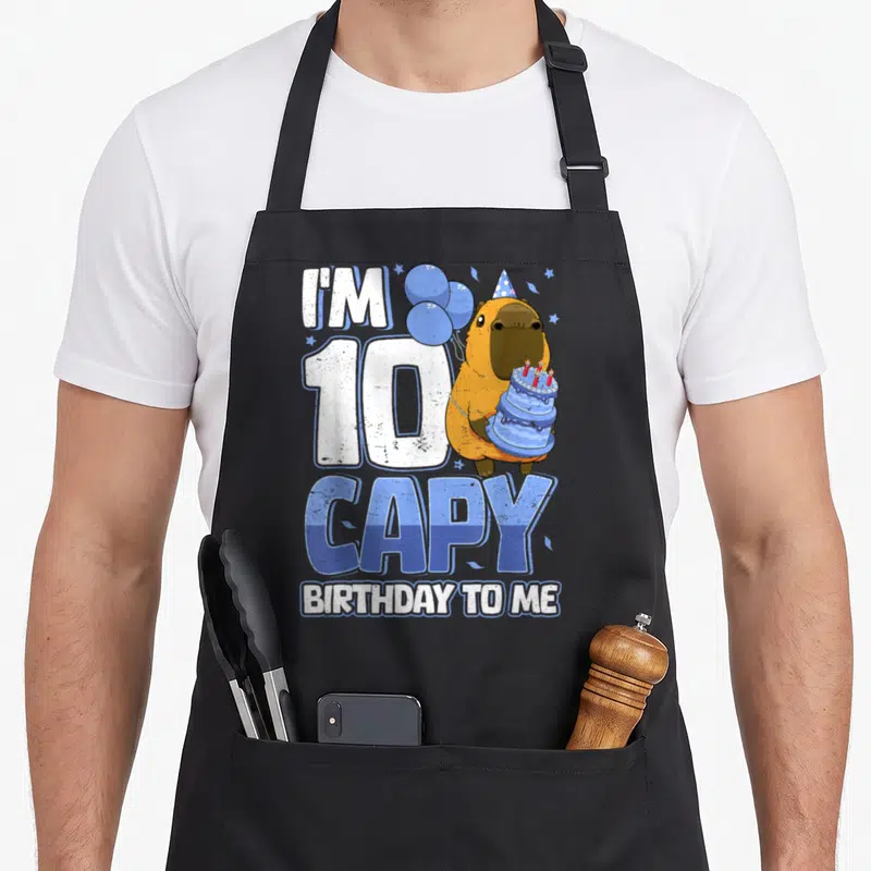 10th capybara birthday apron - Capybara in party hat holding blue cake with balloons and text I'M 10 CAPY BIRTHDAY TO ME — shown on a black cotton canvas apron against a white background
