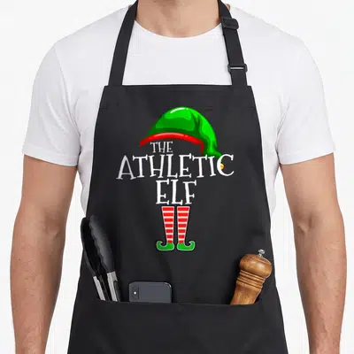 Athletic christmas elf apron - Stacked white text THE ATHLETIC ELF between green hat and red striped legs — shown on a black cotton canvas apron against a white background