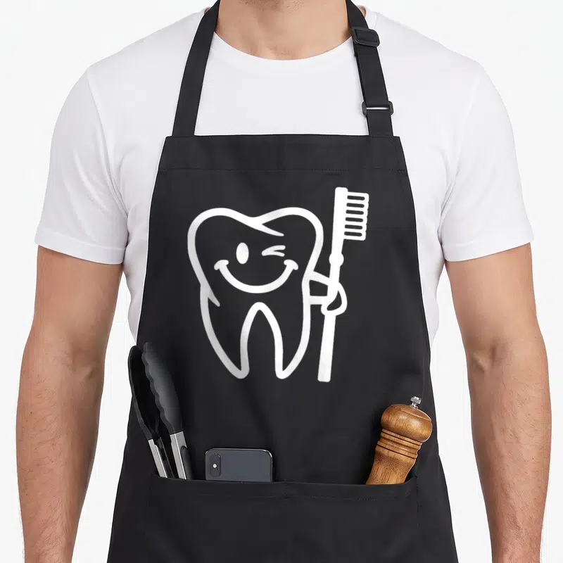 Smiling tooth apron - White cartoon tooth winking and holding a toothbrush — shown on a black cotton canvas apron against a white background