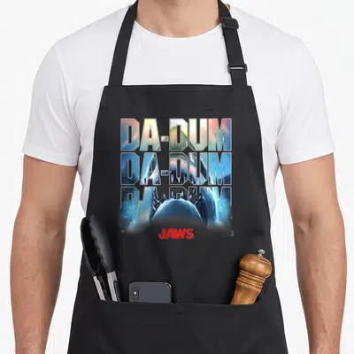 Dum novelty apron - Stacked DA-DUM text containing a shark image above a red JAWS logo — shown on a black cotton canvas apron against a white background