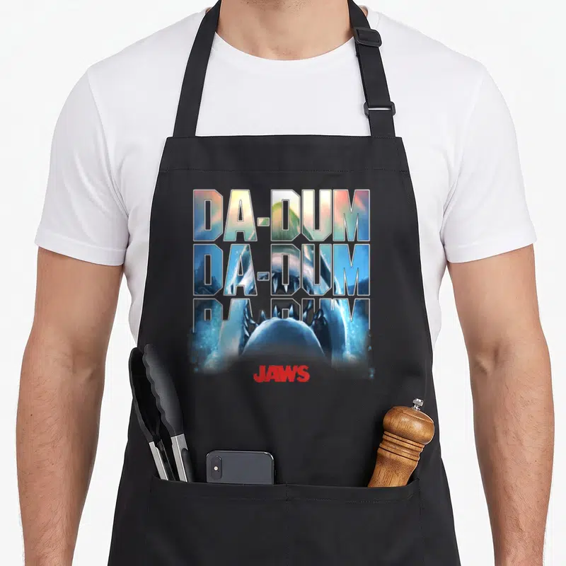 Dum novelty apron - Stacked DA-DUM text containing a shark image above a red JAWS logo — shown on a black cotton canvas apron against a white background