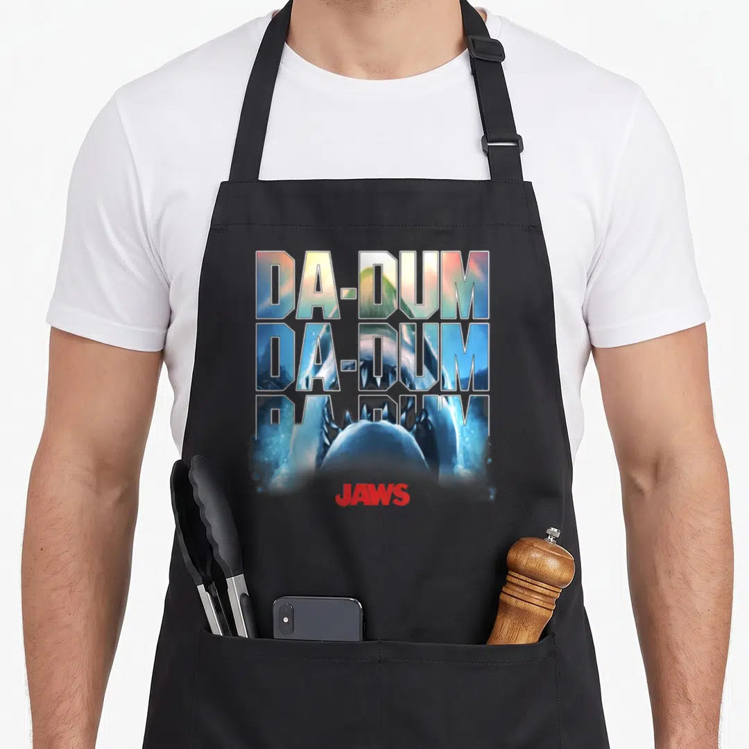 Dum novelty apron - Stacked DA-DUM text containing a shark image above a red JAWS logo — shown on a black cotton canvas apron against a white background