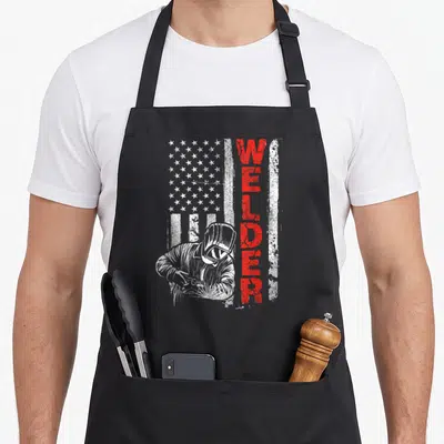 Welder metalworking apron - Distressed USA flag background with vertical red WELDER text and welder in mask illustration — shown on a black cotton canvas apron against a white background