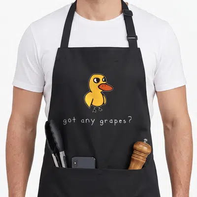 Summer duck apron - Yellow cartoon duck with orange beak and white text reading got any grapes — shown on a black cotton canvas apron against a white background