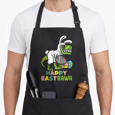 Eastrawr egg apron - Green dinosaur in white bunny suit holding striped egg with HAPPY EASTRAWR text — shown on a black cotton canvas apron against a white background