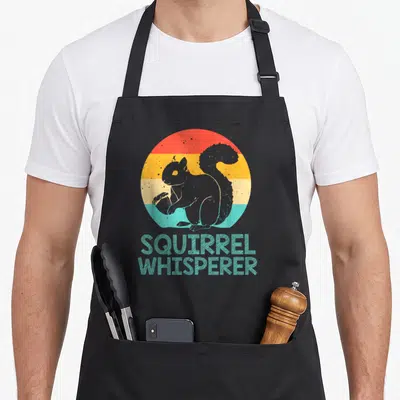 Funny whisperer squirrel aprons - Grey squirrel holding nut on red orange yellow blue striped circle with SQUIRREL — shown on a black cotton canvas apron against a white background
