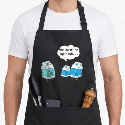 Must spanish aprons - Three cartoon milk cartons, soy milk label, speech bubble he must be spanish — shown on a black cotton canvas apron against a white background