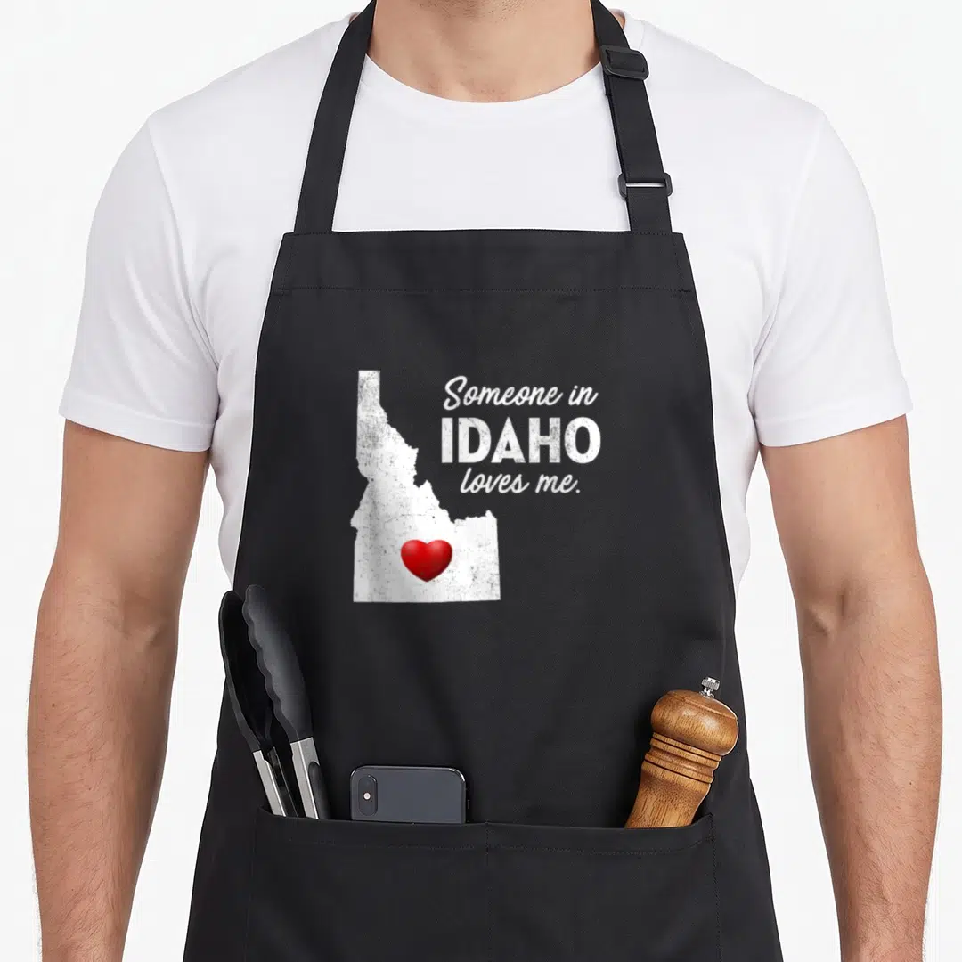 Someone idaho apron - White distressed Idaho map with red heart and text Someone in IDAHO loves me — shown on a black cotton canvas apron against a white background