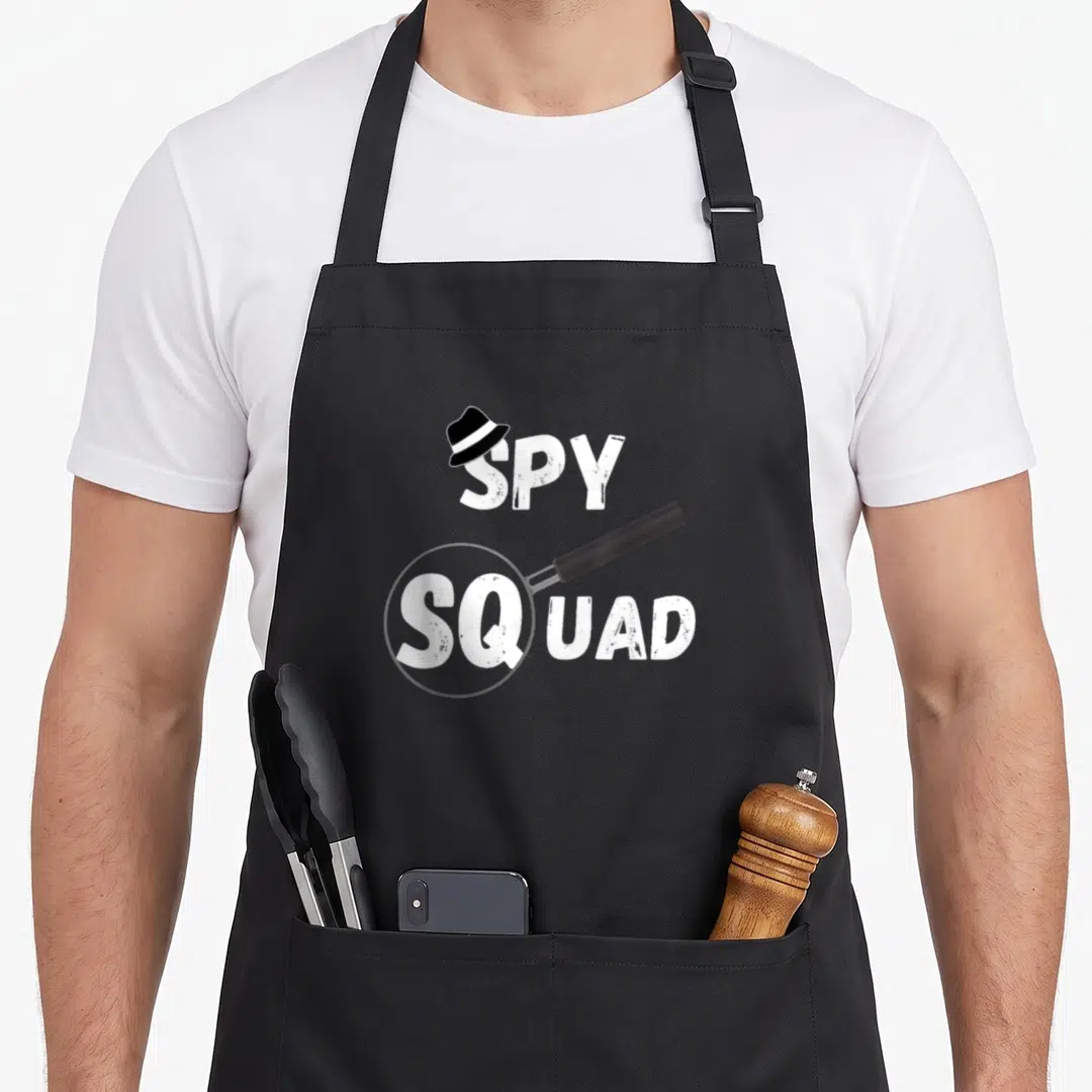 Spy crew apron - White SPY SQUAD text with black fedora on S and magnifying glass around SQ on grey background — shown on a black cotton canvas apron against a white background