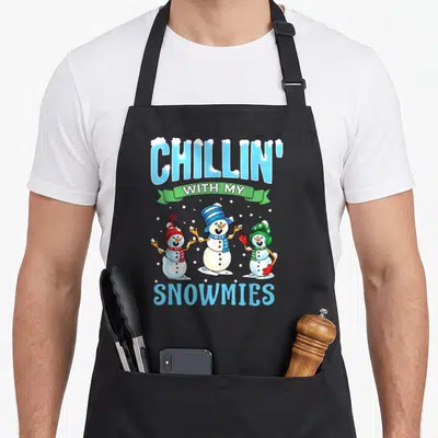 Chillin snowman apron - Three cartoon snowmen under CHILLIN WITH MY SNOWMIES text in blue and green — shown on a black cotton canvas apron against a white background