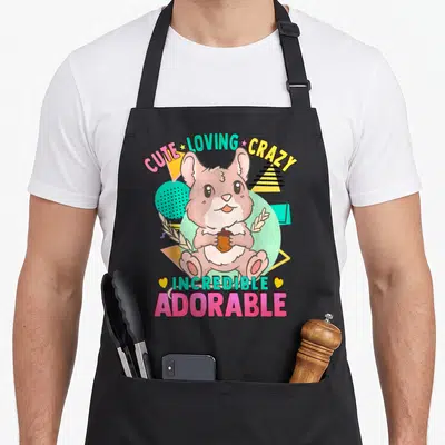 Field kawaii apron - Cartoon hamster holding acorn with CUTE LOVING CRAZY INCREDIBLE ADORABLE text in pink teal yellow — shown on a black cotton canvas apron against a white background